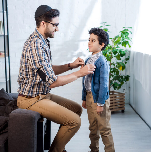A Jewish man wearing a yarmulke adjusts the jacket of a young boy wearing a yarmulke indoors.